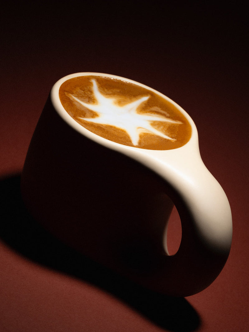 Glamour shot of an milky coffee beverage in a cream chunky ceramic cup. The latte art has the shape of a start with 7 arms. The background is brown.