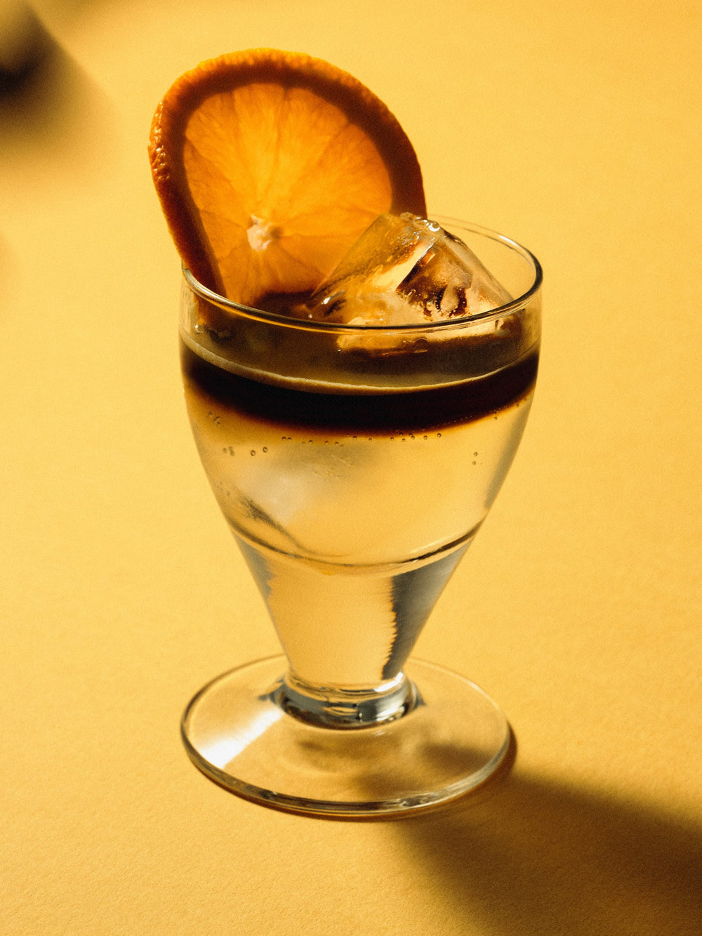 Glass of iced orange marmalade cocktail topped with a dark syrup layer and garnished with a slice of orange. Set on a warm yellow surface with dramatic lighting.