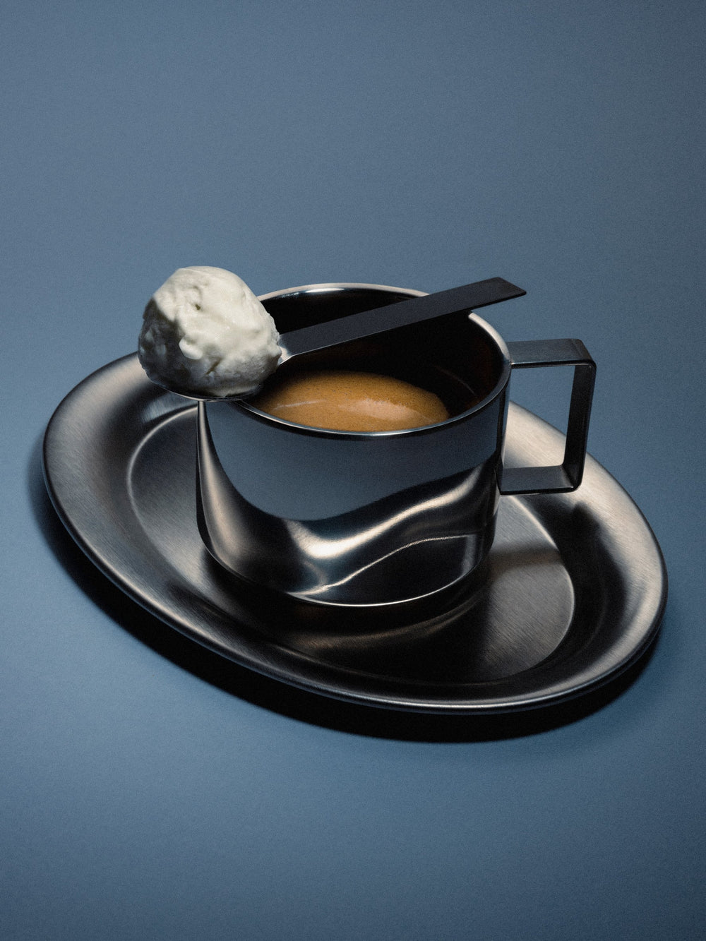 Espresso set in chrome with a hazy light reflexion. Expresso is visible in the cup, and topped with a spoon of whipped cream. The background is blue. 